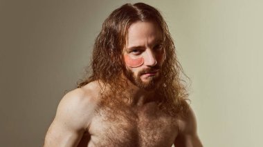 Uzun dalgalı saçlı ve göz altı bantlı sakallı adam, sıcak tonlu arka plana karşı modern erkek derisini temsil eden yoğun bakışlar atıyor. Erkek güzelliği kavramı, cilt bakım ürünleri