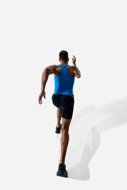 Arka planda mavi formalı ve siyah şortlu beyaz stüdyo arka planında yalnız koşan erkek koşucunun görüntüsü var. Spor, rekabet, dayanıklılık, atletizm, spor yaşam tarzı kavramı