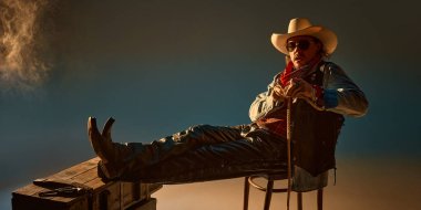 Kovboy olarak vahşi bir adam, kendine güvenen pozuyla sandalyede oturuyor, ahşap göğse yaslanıyor, geniş kenarlı şapka, deri pantolon, kot ceket ve ip tutuyor. Vahşi batı kavramı, Amerikan kültürü.