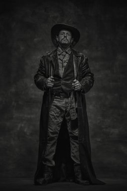 Vahşi, ciddi kovboy şapkalı, uzun deri ceketli, gömlekli, kot pantolonlu, beline ekose gömlek takmış, ileriye bakıyor. Siyah beyaz portre. Amerikan kültürü, tarihi, vahşi batı konsepti