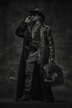 Vahşi bir adamın siyah beyaz portresi, kovboy şapkalı şerif, uzun deri ceketli, gömleğinin üstünde yelek, büyük deri çanta tutan, sigara içen, karanlık arka plana bakan. Amerikan kültürü kavramı