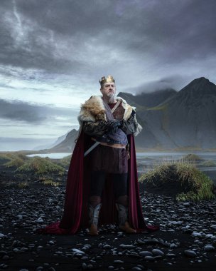 Ortaçağ kralı, kürklü pelerin ve zırhlı, elinde kılıç, bulutlu gökyüzünün altında sisli dağlarla kayalık kıyılarda duruyor. Tarih kavramı, kraliyet, güç, fantezi.