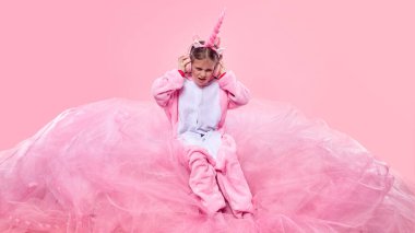 Pembe, sıcak, tek boynuzlu at tulumu giymiş, yumuşak kumaşın üzerinde oturan ve pembe arka planda kulaklıkla müzik dinleyen bir çocuk. Çocukluk kavramı, fantezi, hayaller, hayal gücü, duygular