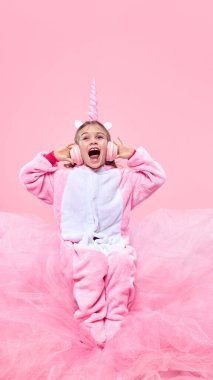 Pembe tulumlu tek boynuzlu at kostümü giymiş çocuk yumuşak kumaş üzerinde oturuyor ve pembe arka planda kulaklıkla müzik dinliyor. Çocukluk kavramı, fantezi, rüyalar, hayal gücü