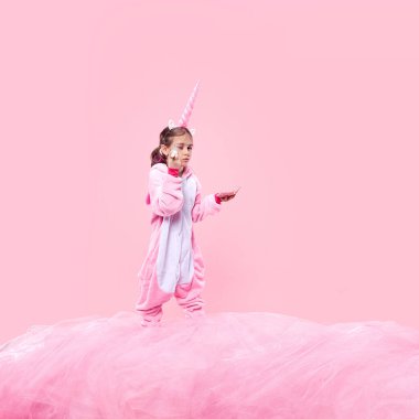 Pembe tek boynuzlu at tulumlu bir kız. Pembe tüylü pembe kumaşın üzerinde duruyor. Küçük bir ayna tutuyor ve pembe stüdyo arka planına fırçayla makyaj yapıyor. Çocukluk kavramı, fantezi, rüyalar, hayal gücü
