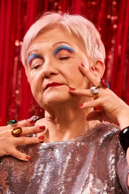 Yaşlı kadın portresi koyu mavi göz farı, kırmızı dudaklar, ve lüks yüzükler, kırmızı arka planda zarif bir yüze dokunuyor. Güzellik güveni. Pop sanat fotoğrafçılığı. Retro cazibe.