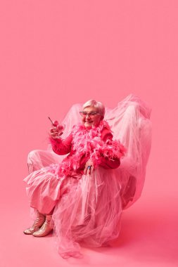 Pembe sandalyede oturan, elinde pembe dondurmalı cam tutan abartılı pembe elbiseli şık, yaşlı bir kadın. Cesur kendini ifade etme. Pop sanat fotoğrafçılığı. Eksantrik yaşam tarzı ve tuhaf moda kavramı