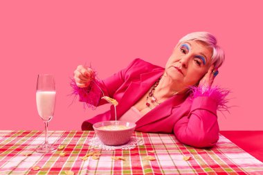 Canlı pembe kıyafetli yaşlı bir kadın elinde mısır gevreğiyle zarif bir kaşık tutuyor, elinde dinleniyor, pembe arka plana karşı sakin bir ifadeyle bakıyor. Yemek ve sanat fotoğrafçılığı. Moda kavramı
