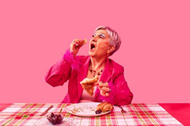 Pembe takım elbiseli yaşlı kadın pembe stüdyo arka planında kruvasan yiyor. Canlı kahvaltı sahnesi. Yemek ve sanat fotoğrafçılığı. Moda, yemek, eğlence ve keyif anlayışı