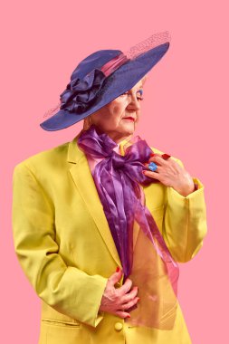 Sarı ceketli yaşlı kadın geniş kenarlı mavi şapka takıyor pembe arka planda poz verirken saf mor eşarbı ayarlıyor. Pop sanat fotoğrafçılığı. Yaşlı yaşam tarzı, moda kavramı