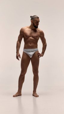 Beyaz iç çamaşırlı, kaslı bir adamın beyaz stüdyo arka planında tanımlanmış fiziğini gösteren tam boy portresi. Erkek güzelliği ve vücut bakımı kavramı.