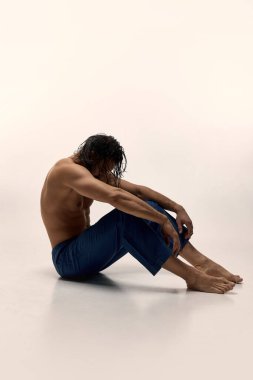 Gömleksiz, kaslı üst vücudu olan, bol pantolonlu, beyaz stüdyo arka planına bakan bir adam. Kişisel bakım, erkek güzelliği, sağlık, ruh sağlığı kavramı