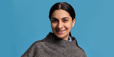 Koyu renk saçlı, gri yün kazaklı ve mavi gömlekli gülümseyen genç bir kadın. Mavi arka planda kameraya bakıyor. Neşe kavramı, pozitiflik, insani duygular, yaşam tarzı.