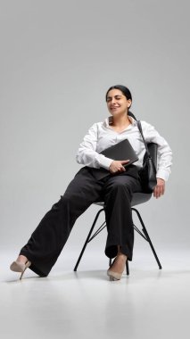 Koyu saçlı, beyaz tişörtlü, siyah pantolonlu, dizüstü bilgisayarlı sandalyede oturan ve gri arka planda gülümseyen rahat bir genç kadın portresi. İş-yaşam dengesi kavramı, güven