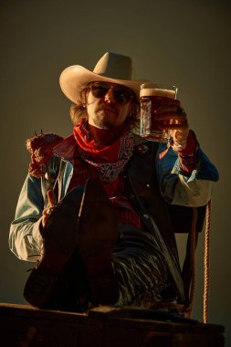 Yakışıklı adam, deri pantolonlu kovboy, bandana, kot ceket, şişte ızgara et ve bira tutuyor, arkaya yaslanıyor. Yiyecek ve içecek kavramı, kırsal bar, vahşi batı tarzı.
