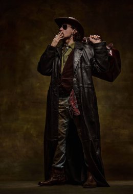 Kovboy şapkalı, güneş gözlüklü, deri ceketli, deri çantalı, koyu renk stüdyo arka planında sigara içen yakışıklı bir adamın tam boy portresi. Amerikan kültürü, tarihi, vahşi batı konsepti