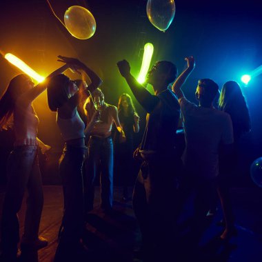 Neşeli bir grup genç arkadaş dans ediyor, neon ışıklı partinin tadını çıkarıyorlar, kahkaha ve enerji dolu. Renkli ışıklar, gençliğin tasasız neşesini ve bağlantıyı yansıtan neşeli bir atmosfer yaratır..