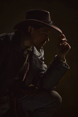 Acımasız düşünceli bir adamın portresi, deri ceket giymiş kovboy ve karanlık arka plana karşı sessiz güç ve bilgelik saçan bir şapka. Dayanıklılık kavramı, sınır hayatta kalma, batı tarihi.