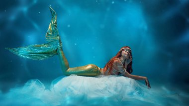 Güzel kızıl saçlı kadın, narin kumaşın üzerinde yatan deniz kızı, düşüncelerde kaybolmuş, parlak mavi yansımalarla çevrili. Fantezi, gizem, huzurlu rüyalar ve derin deniz büyüsü kavramı..