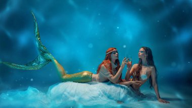 İki zarif genç kadın, şampanyanın tadını çıkarıyorlar. Etrafı mavi ışıkla çevrili. Derin okyanus kutlaması. Huzur, fantezi, ruhani cazibe ve sualtı zarafeti kavramı..