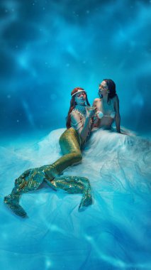 Derin mavi okyanus ışığında iki deniz kızı inciler ve parlak kuyruklarla süslenmiş, samimi bir anı paylaşıyorlar. Fantezi kavramı, efsanevi yaratıklar, derin deniz sihri, dostluk ve ruhani güzellik..