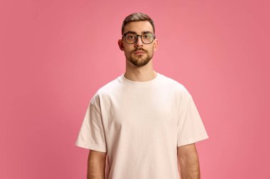 Beyaz tişörtlü ve gözlüklü genç adam pembe arka planda ciddi bir ifadeyle poz veriyor. İnsan duyguları, yaşam tarzı, basitlik, özgüven, moda, erkeklik kavramı.