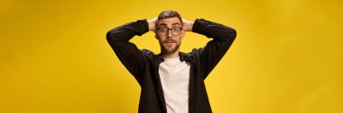 Siyah beyaz kıyafetli, şok olmuş ve heyecanlı genç bir adamın portresi sarı stüdyo arka planına karşı gözleri açık bir şekilde başının arkasında el ele tutuşuyor. İnsan duyguları kavramı, yaşam tarzı, reklamlar