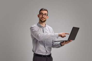 Genç adam kendine güveni olan, modern ofis kıyafetleri içinde duran bir dizüstü bilgisayar ekranını işaret ediyor. Dijital yaşam tarzı, odaklanma, eğitim, çalışma süreci, anlamlı insan etkileşimi, iş