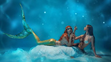 Altın kuyruklu güzel bir denizkızı kız arkadaşlarıyla birlikte yüzen kumaşın üzerinde uzanmış, etrafında parlak mavi yansımalarla çevrili şampanya içiyor. Gizem, fantezi, mitoloji ve okyanus güzelliği kavramı.