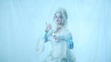 Kraliyet kostümlü zarif genç bayan açık mavi arka planda kaşıkla yumurta kırıyor. Dönem, güzellik, tarih, sanat, yaratıcılık karşılaştırması.