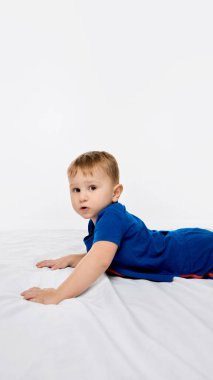 Karnında beyaz çarşaflarla yatakta uzanmış beyaz arka plana meraklı meraklı bakan bir çocuk. Erken çocukluk kavramı, büyüme, sağlık, hareket, bilişsel uyarı, öğrenme, oyun.