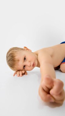 Küçük çocuk yere uzanmış, eli kameraya doğru uzanmış ve beyaz arka planda meraklı meraklı bakıyor. Çocukluk kavramı, büyüme, keşif, gelişme, oyun, duygu, sağlık.
