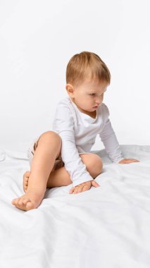 Beyaz stüdyo geçmişine karşı meraklı bakışlarla yatakta oturan odaklanmış küçük bir çocuk. Çocukluk kavramı, öğrenme, duygu, sağlık, büyüme, davranış, gelişme.