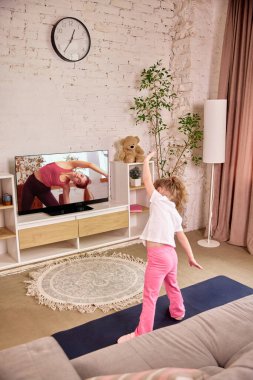 TV 'de beden eğitimi hocası seyreden bir kız, sabah antrenmanı sırasında kollarını kaldıran, evde antrenman yapan bir kız. Çocukluk kavramı, çevrimiçi eğitim, uzaktan eğitim