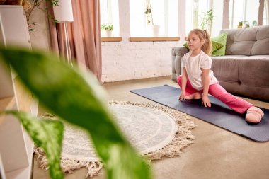 Odaklanmış kız, bir bacağı yandan öbür yana uzanmış yeşil bitkilerle güneşli oturma odasında antrenman yapıyor. Çocukluk kavramı, çevrimiçi eğitim, uzaktan eğitim