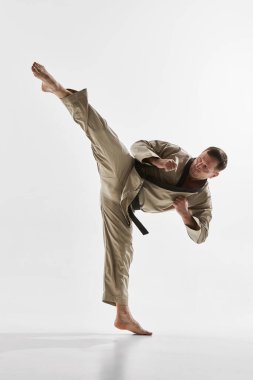 Yetişkin erkek tekvando dövüşçüsü bej dobok giyip beyaz arka planda yüksek tekme atıyor. Dövüş sporu kavramı, dövüş sanatları, güç, denge, kontrol, hareket, disiplin, güç