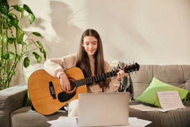 Çok güzel. Modern, şık oturma odasındaki kanepede oturan gülümseyen genç kız sanal müzik dersi sırasında dizüstü bilgisayara bakarken gitar çalıyor. Evde eğitim, eğitim, özel öğretmen kavramı