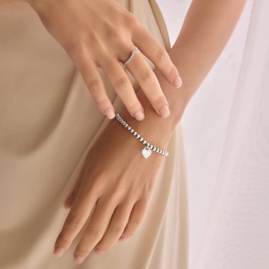 Gümüş aksesuar takan hassas kadın ellerinin yakın plan görüntüsü. Hafif bulanık arkaplanlı hafif kumaşa karşı kalp çekiciliğine sahip narin bir yüzük ve bilezik. Mücevher kavramı, lüks.