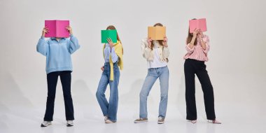 Yüzleri kaplayan renkli kitaplarla poz veren bir grup öğrenci, modern sınıfta beyaz arka planda gelişigüzel duruyorlar. Eğitim kavramı, üniversite, eğlenceli öğrenme, gençlik kültürü