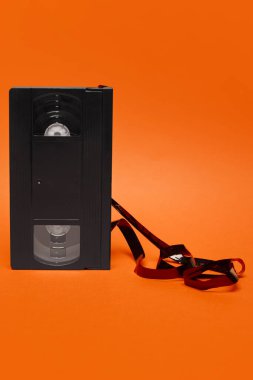 Turuncu arka planda çözülmüş manyetik kurdeleli siyah VHS bandı, analog nostaljiyi ve pop kültürünün bozulmasını sembolize ediyor. Retro estetik, vintage, pop sanat, tasarım kavramı.