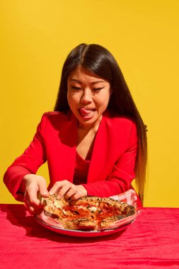 Lezzetli pizza yemek için heyecanlanan genç Asyalı kadın, parlak stüdyo arka planına karşı tabaktan dilimler alıyor. Yemek kültürü kavramı, pop sanat fotoğrafçılığı, insan duyguları.