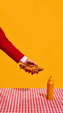 Erkeğin dikey görüntüsü, sarı stüdyo arkaplanına karşı hardal şişesiyle kareli masa örtüsünün üstünde sosisli sandviç tutuyor. Fast food estetiği, pop sanat fotoğrafçılığı, restoran menüsü