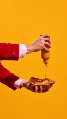 Sosislinin üzerine koyu sarı arka planda hardal döken eller. Poster, afiş, reklam. Fast food tanıtımı. Fast food estetiği kavramı, pop sanat fotoğrafçılığı.