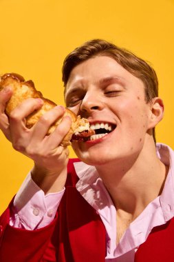 Kırmızı elbiseli yakışıklı genç adamın yakından çekilmiş portresi. Cesur sarı arka planda tutkulu bir şekilde lezzetli sosisli yiyor. Fast food estetiği kavramı, pop sanat fotoğrafçılığı, duygular