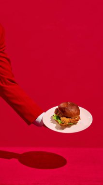 Kırmızı takım elbiseli, elinde tavuk burgerli beyaz bir oyuncuyla canlı kırmızı stüdyo arka planına bakan bir erkeğin dikey görüntüsü. Fast food estetiği kavramı, pop sanat fotoğrafçılığı. Boşluğu kopyala