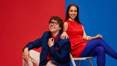 Gülümseyen genç adam ve kadın, kırmızı ve mavi arkaplan karşısında eşleşen kıyafetler içinde birlikte oturuyorlar. İnsan duyguları kavramı, mutluluk, neşe, ilişki, yaşam tarzı