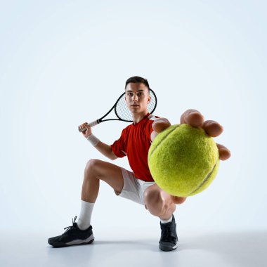 Kırmızı ve mavi üniformalı genç adam tenis topu ve raketle poz veriyor, beyaz stüdyo arka planına güçlü bir atış için hazır. Spor, oyun, turnuva, pratik, gençlik kavramı