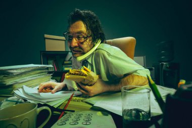 Ofisteki adam gergin bir şekilde belgeleri kontrol ediyor, sandviçi tutuyor, telefonda konuşuyor, stres gösteriyor, absürd, iş yükü, tükenmişlik ve çoklu görev. Eski ofis kavramı, iş hayatı, son teslim tarihi