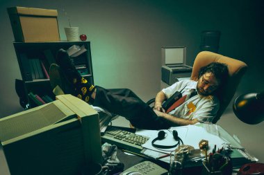 Gözlüklü bir adam masa başında bacakları kağıt ve bilgisayarla çevrili bir şekilde uyuyakalır. Bunaltıcı bir çalışma ve dinlenme ihtiyacı. Bitkinlik, retro ofis ve yorgunluk kavramı.