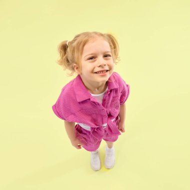 Pembe gömlek ve şort giyen at kuyruklu gülümseyen kız, sarı arka planda neşeyle poz veriyor. Mutluluk ve rahatlık. Çocukluk kavramı, yaz tatili, aktif yaşam tarzı, eğlence